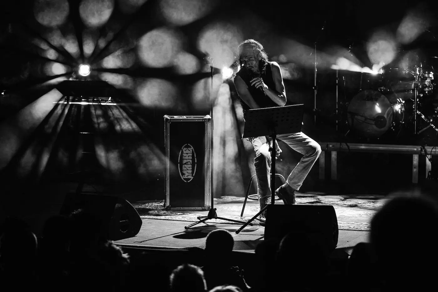 Male singer on stage with microphone, music stand, and dramatic stage lights, performing to an audience in a concert hall (black and white).