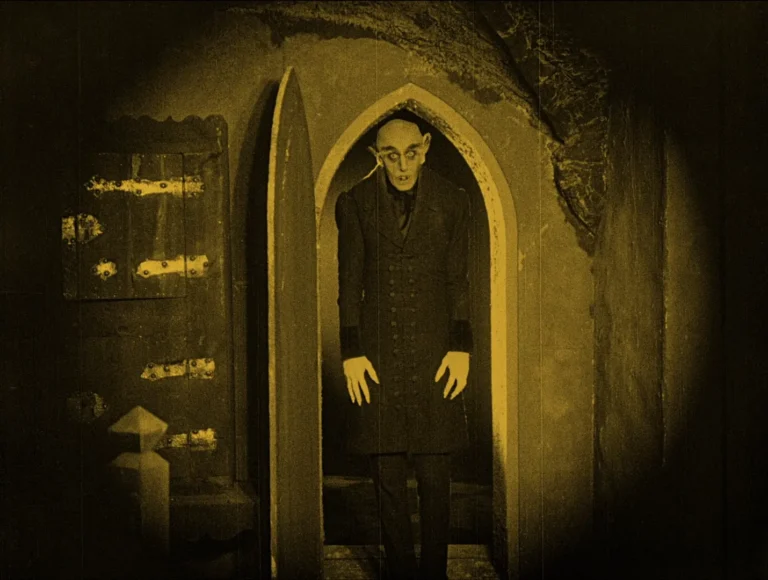 Dim, sepia-toned scene: a pale man in a dark coat stands in a Gothic arched doorway, reaching toward the camera.