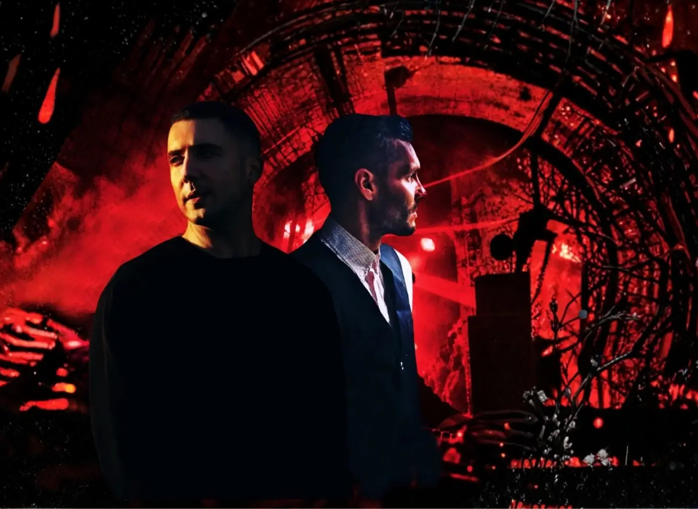 Two men stand back-to-back on a red-lit stage with smoky atmosphere and an industrial backdrop.